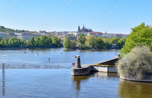 A scenic view of Prague across the Vltava River, with Prague Castle and St. Vitus Cathedral visible on the hillside, and a river lock and navigation beacon in the foreground.