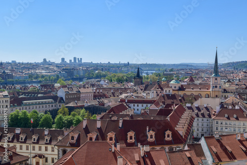 A panoramic view of a Prague city featuring red-tiled rooftops, church spires, and classical architecture. In the distance, modern skyscrapers contrast with the city's well-preserved old town