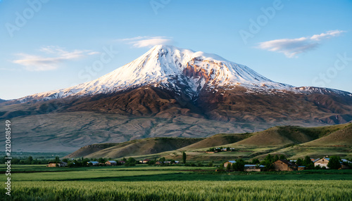 Majestic snow-capped volcano rising over green valley. Dramatic mountain peak with glacier under clear blue sky for travel and nature projects.