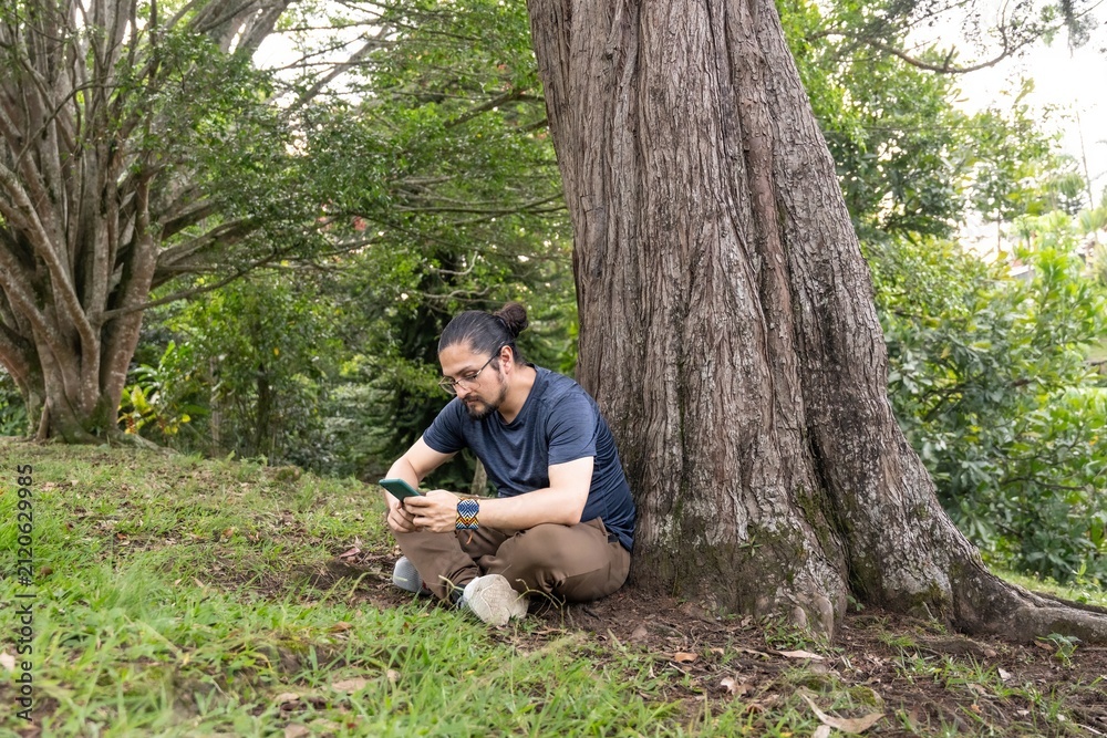 Fototapeta premium Man using smartphone sitting under tree trunk in green park
