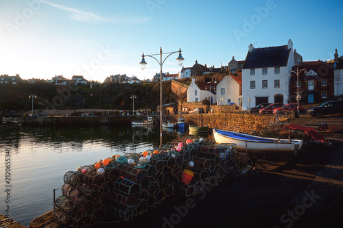 Quiet waterfront harbor with fishing boats, lobster pots, stone walls, and village houses in warm evening light under a clear sky. Crail, Scotland