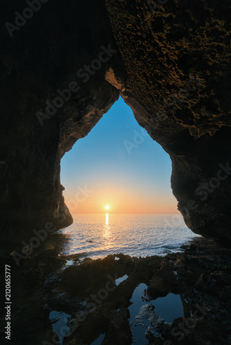 Rocky cave opening frames a calm dawn over the sea with reflections in tidal pools on the Sardinian coast.