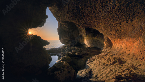 Dark rocky cave frames a golden sunrise over the calm sea along the Golfo di Orosei coast.