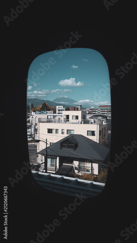 Snow-capped Mount Fuji rises above rooftops and clouds, framed by the oval window of an train.