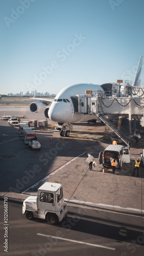 A wide-body passenger airplane parked at an airport jet bridge, surrounded by ground crew and service vehicles loading cargo under a clear sky.