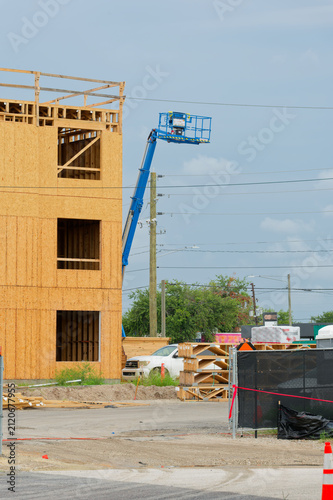 A mid-rise building under construction featuring oriented strand board wood framing, a blue boom lift raised beside the structure, stacked lumber, safety fencing and utility poles with overhead power 