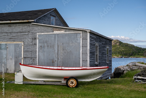 Obraz na plátně A small red and white fishing boat with bright red trim on a trailer sitting on grass in front of a grey, weathered boathouse