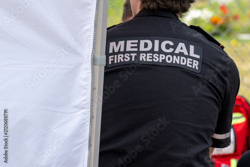 A close-up of a Caucasian male emergency health medical first responder or paramedic wearing a black uniform with grey lettering. The ambulance attendant is wearing a short-sleeve shirt with lettering