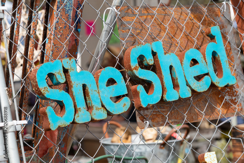 A teal blue colored tin sign, she shed, hanging outdoors on a galvanized metal garden fence. The rustic tinplate decorative wall hanging has spots of rust on the 3D she shed rustic signage. 