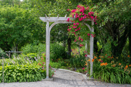 An outdoor wooden flat-top shaped archway or arbour surrounded by a lush green garden.  The park features birch trees, climbing red roses, orange lily flowers, and vibrant green shrubs in a garden.