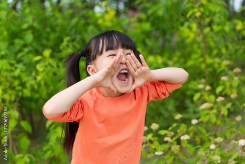 A little girl in an orange shirt stood outdoors, raising her hands, opening her mouth wide, and shouting loudly.