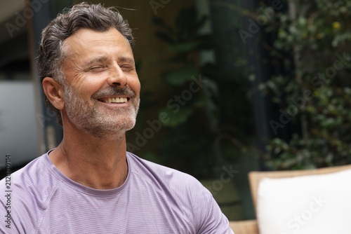 Middle-aged man smiling with eyes closed, sitting on patio chair wearing purple athletic shirt