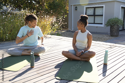 African American couple sitting on green yoga mats on wooden deck with teal water bottles, smiling