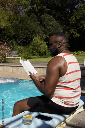 African American man in swim trunks sitting and reading pamphlet at pool with phone, lemon glass