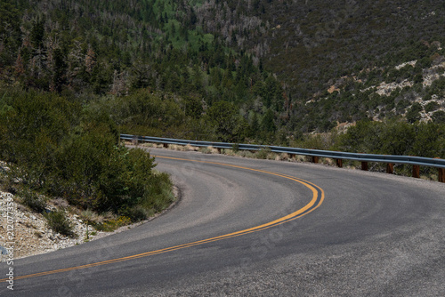 Empty two-lane road curves sharply through a forested mountain canyon, lined with a metal guardrail and dense green trees and shrubs. Steep rocky slopes rise on either side.