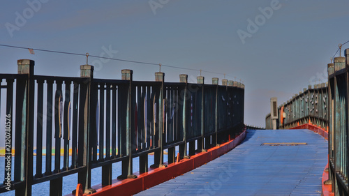 Bild auf Leinwand Curved Blue Pedestrian Bridge Over Water with Metal Railings and Clear Sky