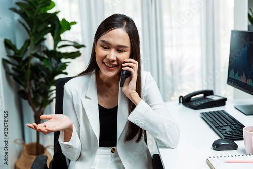 Obraz na plátne Smiling young beautiful Asian businesswoman calling to broker on smartphone to analyze exchange stock market graph on pc screen at modern office
