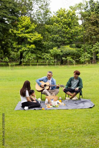GrandFather playing acoustic guitar for child during picnic. Asian family enjoy outdoor picnic with food and drinks in park sunny day, outdoor activitie with daughter father, mother grandfather