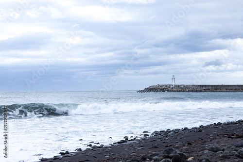 Фотография White lighthouse on a breakwater with crashing winter sea waves on a black pebble beach in Jeju Island, South Korea