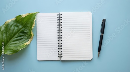Minimalist writing setup with notebook pen and variegated leaf on soft blue background with minimalist workspace with instrument large variegated