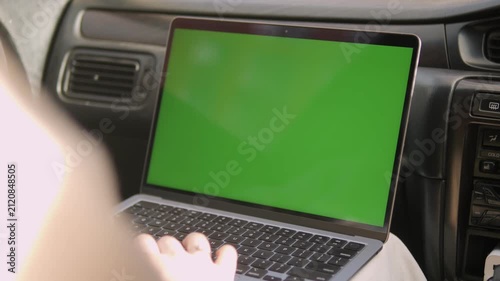 Person uses laptop with green screen inside car. Hands type on keyboard, focused on screen. View shows dashboard and car interior. Good for travel, remote work, or digital content overlays