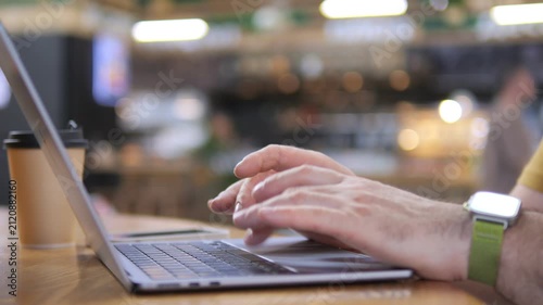 Hands type on laptop in cozy cafe. Warm light glows softly around. Coffee cup rests beside keyboard. Person wears smartwatch and yellow shirt. Scene captures relaxed work or study moment