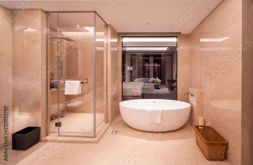A luxurious and modern hotel bathroom featuring a freestanding round white bathtub, a glass-enclosed shower, and elegant beige marble tile walls with a view of the bedroom suite