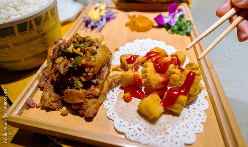 A delicious Asian meal served on a wooden board featuring crispy fried pork, fried tofu with ketchup, and rice, with a hand holding chopsticks ready to eat.