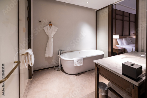 A luxurious and modern hotel bathroom featuring a freestanding round white bathtub, a glass-enclosed shower, and elegant beige marble tile walls with a view of the bedroom suite