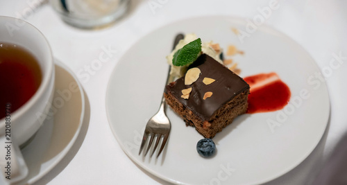 A delicious slice of chocolate cake topped with almonds, cream, mint, and a blueberry, served on a white plate next to a cup of tea, perfect for a gourmet dessert.