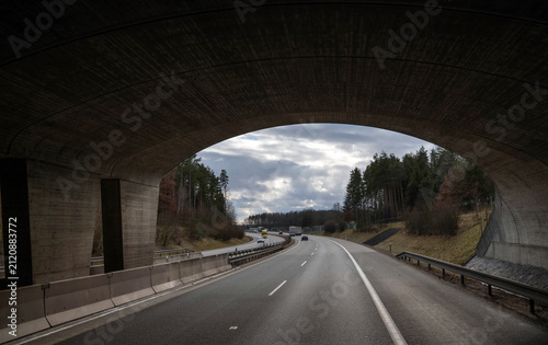 A view from beneath a massive concrete highway bridge framing a road stretching through a landscape with trees and distant vehicles under an overcast sky