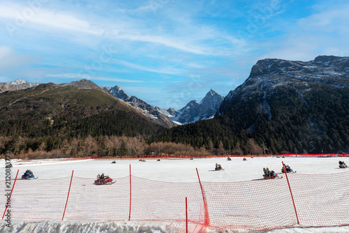 People enjoying snowmobiling on a wide snow-covered field surrounded by majestic snow-capped mountain peaks and evergreen forests under a bright blue sky with light clouds