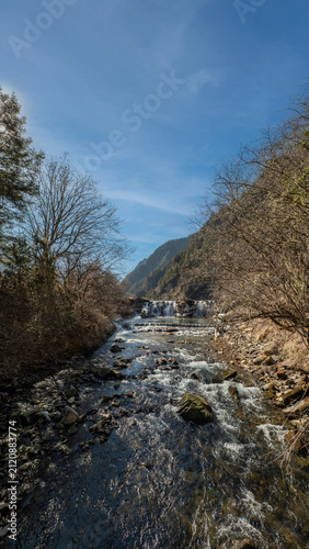 scenic mountain stream with rushing water, rocky rapids, and bare trees along the banks leading to a small waterfall under a clear blue sky on a bright day