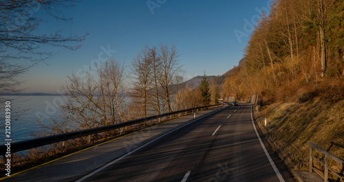 A scenic coastal road stretching alongside a calm lake with bare trees and a forested hill under a clear blue sky on a sunny day during a road trip