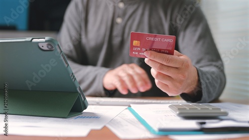 Man holds red credit card, types tablet keyboard, calculates business expenses, manages personal finance, pays bills online, performs electronic commerce transactions inside home workspace office.
