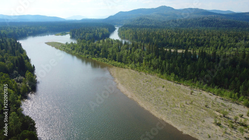 Aerial drone view of wild river flowing through taiga forest surrounded by mountains in summer. Untouched nature landscape with green trees and clean water. Concept of wilderness, remote travel 