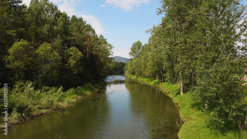 Drone shot of river in taiga surrounded by mountains and dense forest in summer. Scenic aerial view for travel, adventure and exploration concepts. Remote destination with wild untouched nature and be