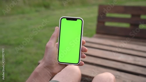 Hand holds smartphone with green screen. Person sits on wooden bench outdoors. Background shows grass and nature. Ideal for app or advertisement mockups
