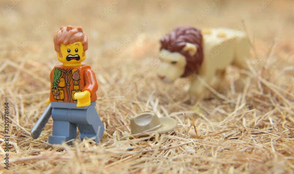 Fototapeta premium Lego minifigure of a ranger with machete is running away from the lion in the natural dried grass field. Editorial illustrative image of popular plastic toy.