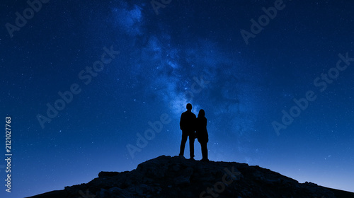 Silhouette of young couple standing on hilltop looking at starry night sky with milky way 