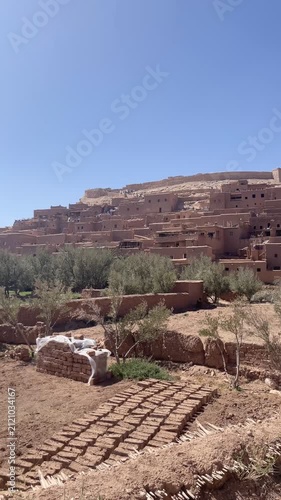 panoramic view of ancient fortified clay ksar village Ait Benhaddou built on hill slope under clear blue sky in Morocco, In the foreground, a new building with stacked adobe bricks