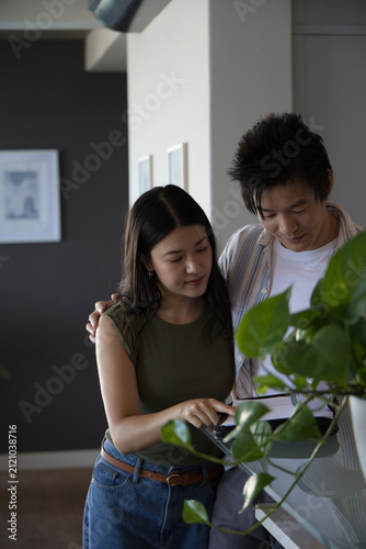 Asian couple standing and leaning over tablet at kitchen counter near potted plant, pointing
