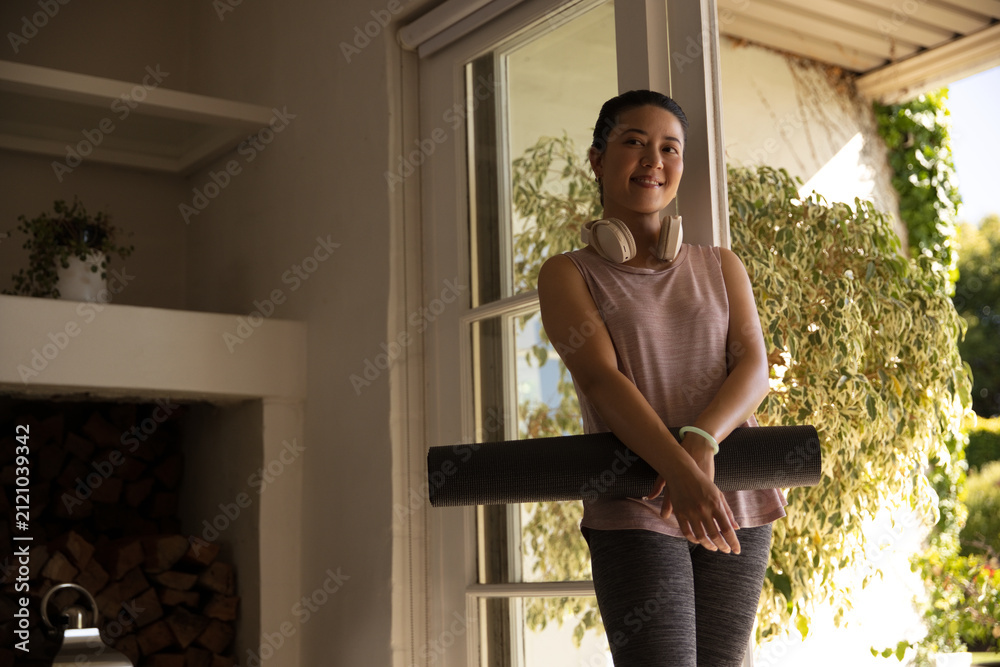 Naklejka premium Adult Asian woman holding yoga mat in home doorway in workout clothes, wearing headphones