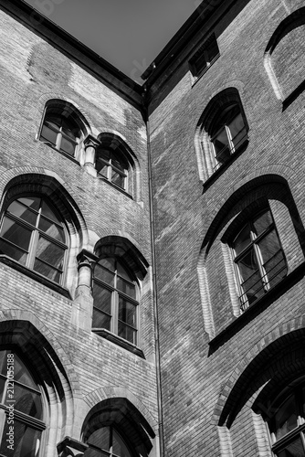 Photography Black and white view of a brick courtyard corner with arched windows and stone mullions, strong sunlight raking across the facades