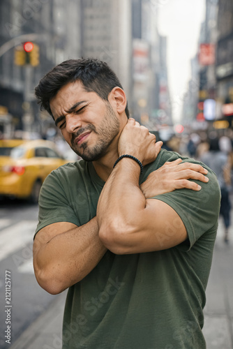 A young man holds his neck in pain while standing on a street. The background shows a busy city. AI generated