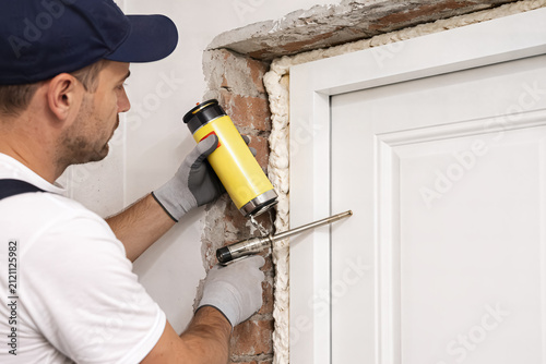 A construction worker seals a door frame with yellow foam. He wears gloves and uses a spray gun tool. AI generated