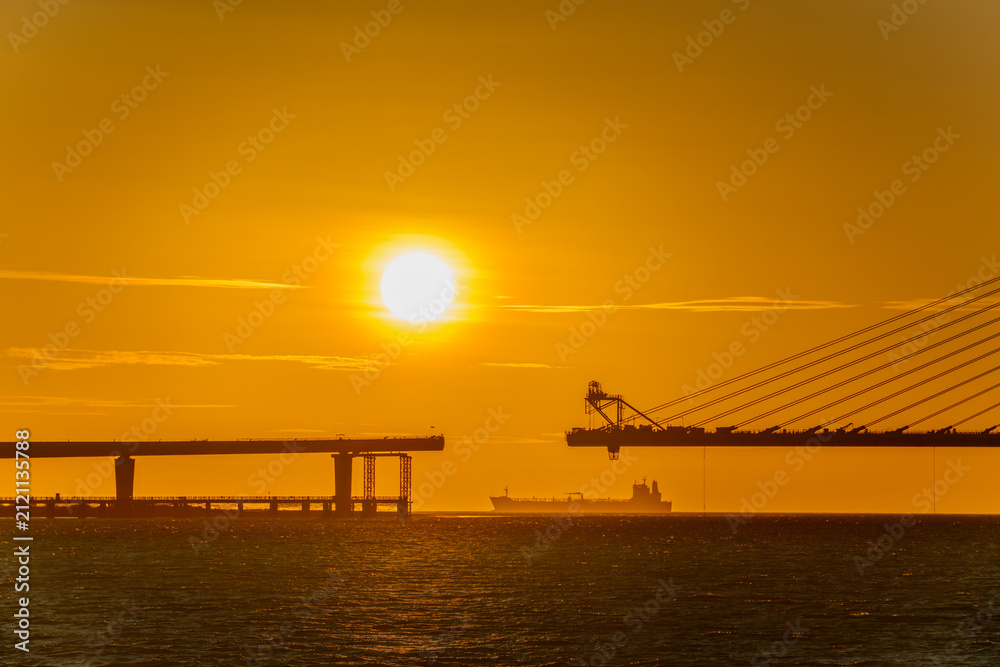 Fototapeta premium A scenic sunset view of the Danjiang Bridge under construction in Tamsui, New Taipei City, Taiwan, with a cargo ship passing by in the distance.