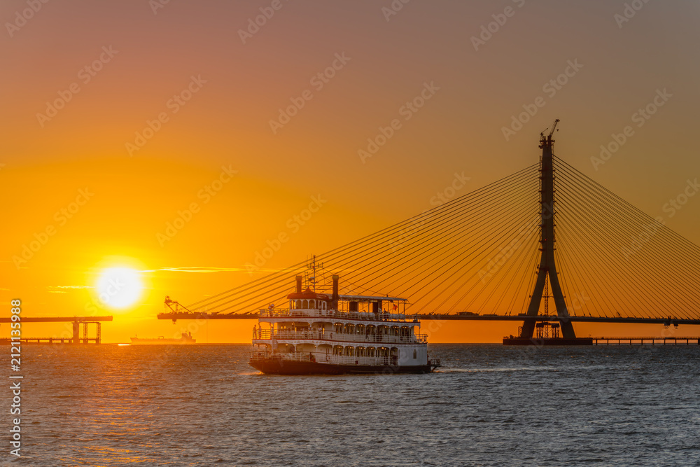 Fototapeta premium A scenic sunset view featuring a passenger ferry boat sailing near the construction site of the Danjiang Bridge in Tamsui, New Taipei City, Taiwan.