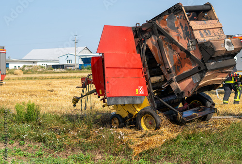 Obraz na plátně Burnt and charred agricultural baler standing on a stubble field after a fire, with firefighters and a fire truck visible in the background under a blue sky on a sunny day