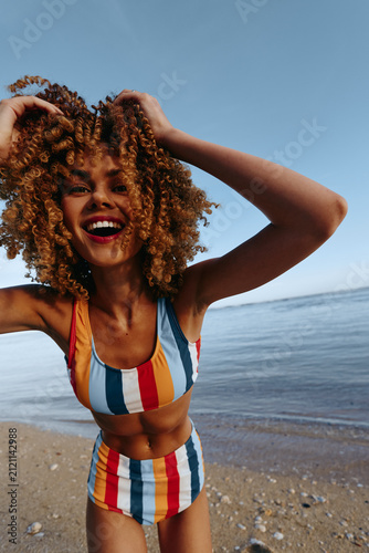 Принти на полотні Joyful woman in striped swimsuit smiles, windblown hair by the beach and calm ocean